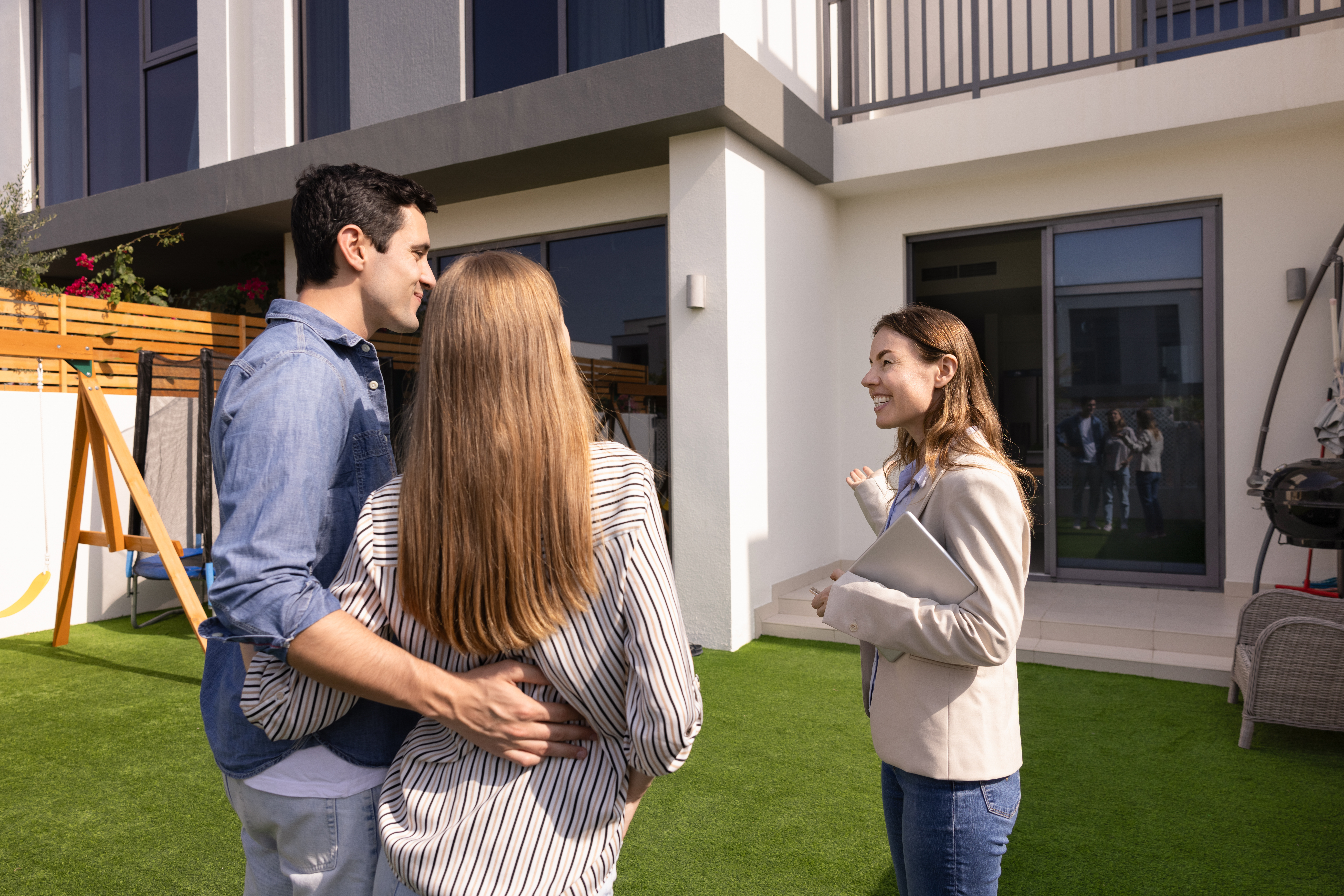 Happy real estate agent woman showing house for sale to young couple of customers, buyers, standing on yard, pointing at exterior, speaking, smiling. Renters meeting with realtor for viewing